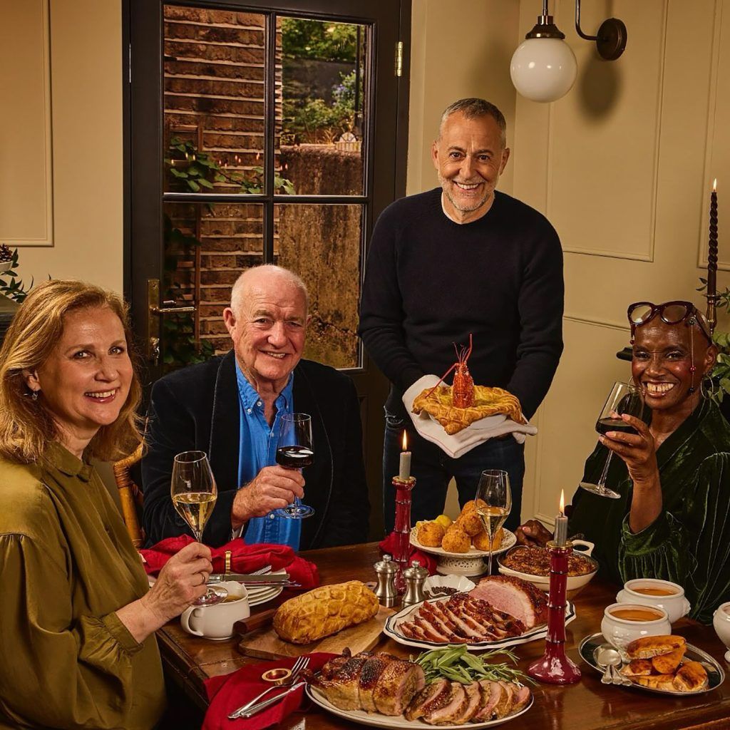 Group portrait of celebrity chefs Michel Roux, Angela Hartnett, Rick Stein and Andi Oliver sat around a dining table drinking wine, with a table full of food, smiling at the camera.