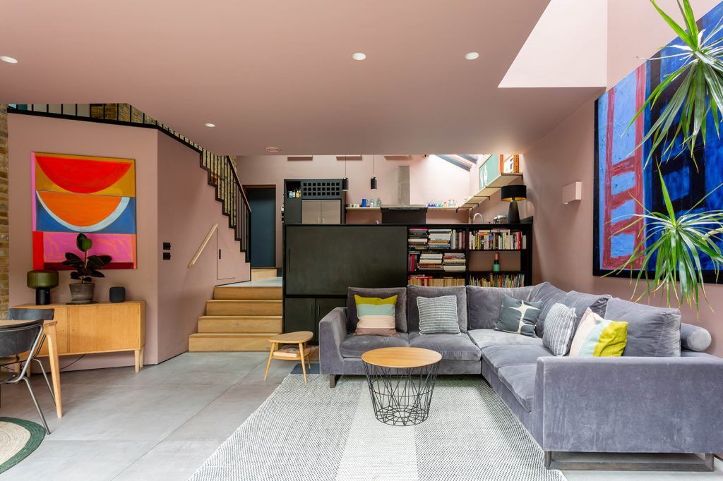 a pink modern living room with grey tiled floor and a grey L shaped sofa. The space is split over two levels with a black media unit and four steps to the kitchen visible behind. 