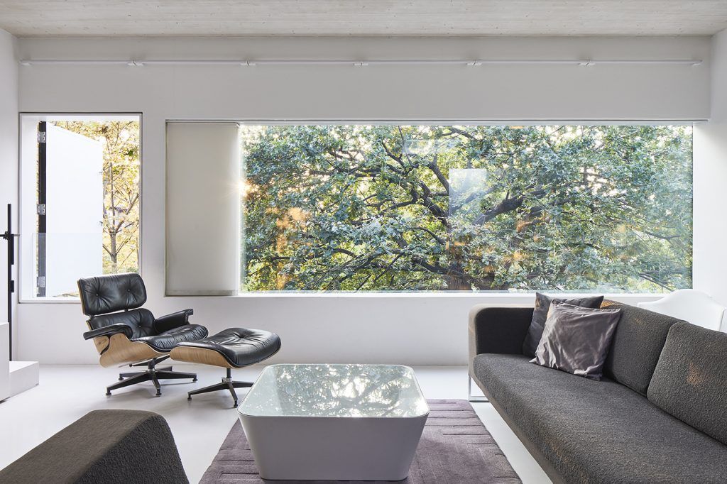 A modern living room with a wide visa view of a tree. The space is white washed with a Eames lounge chair on the left hand side and a grey sofa on the right, in the centre a space age white coffee table with a glass top sits atop a grey rug. 