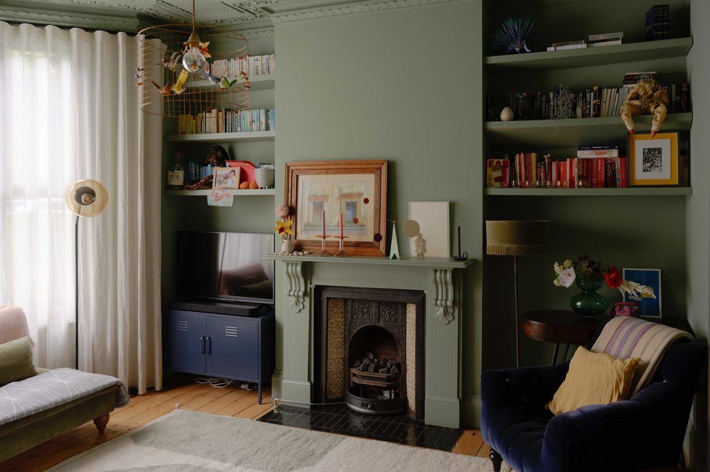 a Victorian living room painted in a soft green colour with built-in bookshelves, a fireplace and warm wooden flooring. There are soft cream curtains, a large rug and a dark navy armchair.