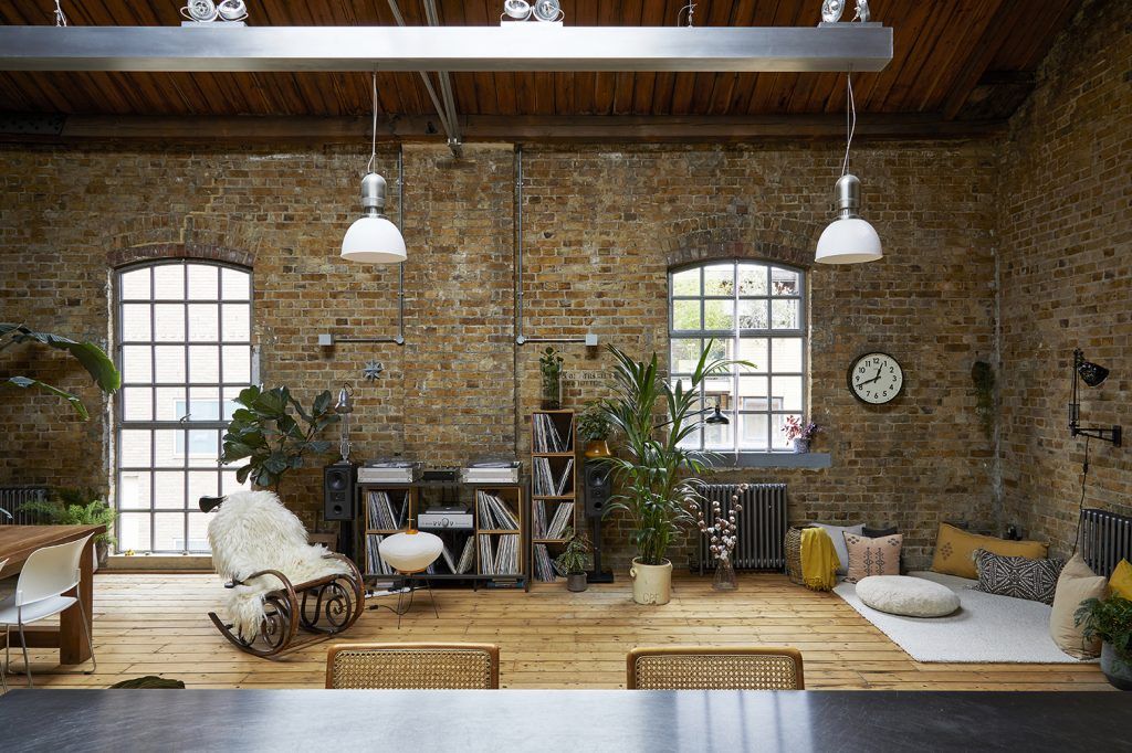 a loft apartment with exposed brick walls, crittal windows and wooden flooring. The space features plants, a record player and record storage, an accent chair and two hanging pendant lights in white. 