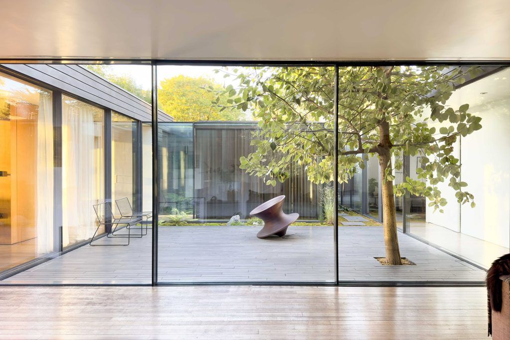 a modern house with a central courtyard, the view is from inside through sliding doors. From all sides glass sliding doors provide a peak into each adjacent space. The courtyard is decked and features a tree to the right hand side, a Spun chair just off centre and then two wire chairs to the left hand side. 