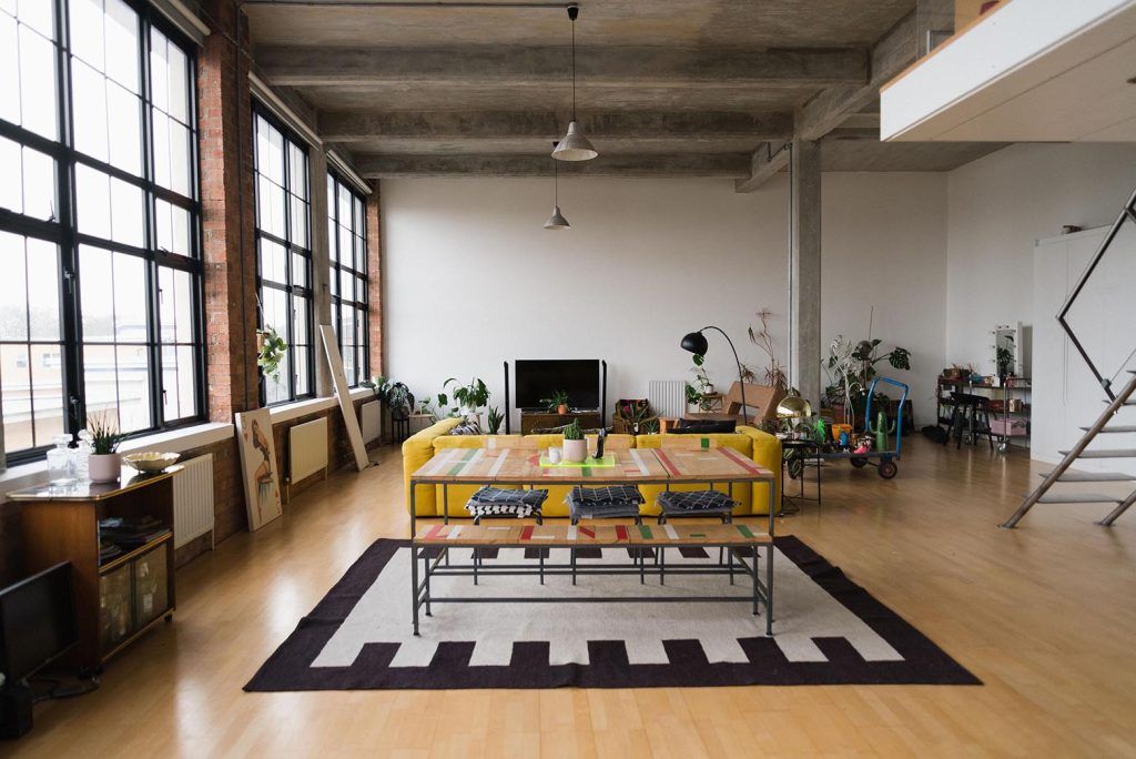 an open-plan loft space with light wooden flooring, concrete ceilings and crittal windows surrounded by exposed brick. The space features a yellow sofa, a table with bench seating set on a cream and balck rug. Other industrial features are seen in the background. 