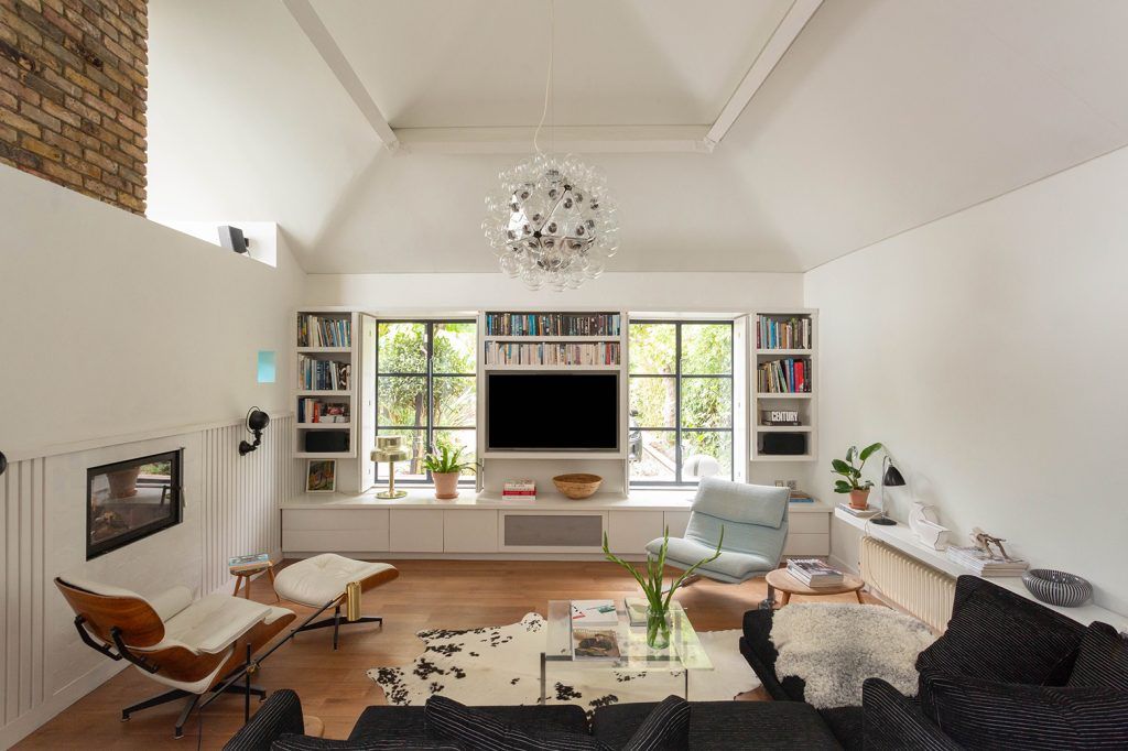 a modern living room with an apex ceiling, light wooden flooring and a large pendant light hanging from the ceiling. In the centre of the image there is a wall-mounted tv, with crittal windows either side and book shelves. There are two mid-century chairs, one an Eames lounge chair, and a large dark sofa. 