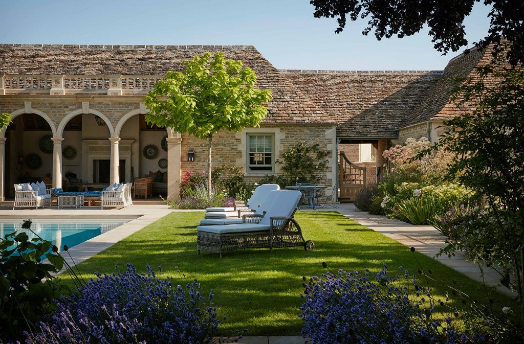 a grand country house with arches and columns, green grass, a swimming pool and sun loungers. Lavender can be seen at the front of the shot and a path runs around the perimeter.   