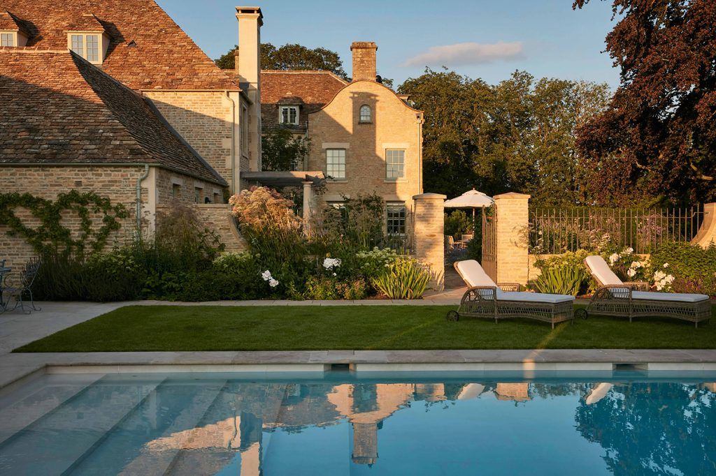 sunset shot of a grand country house with long shadows, green manicured lawn, a swimming pool and two sun loungers. 