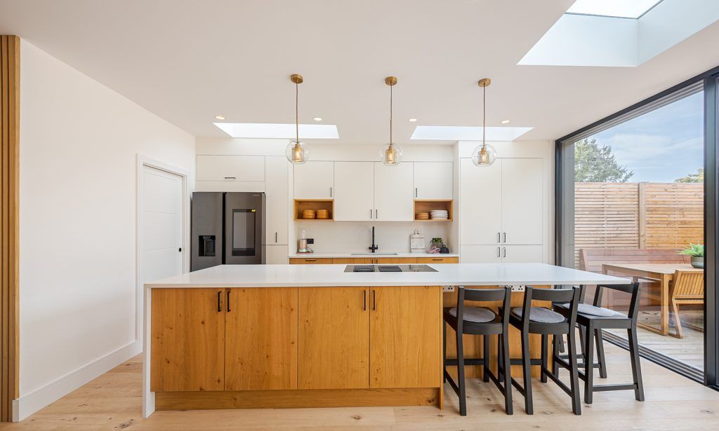 inviting kitchen in a modern extension with skylights, light wooden flooring and sliding doors to a patio. The main feature is a large kitchen island with wooden cupboards and a waterfall white countertop, three black bar stools are to the right hand side and atop the island is an integrated hob. Three pendant lights hang above the island. 