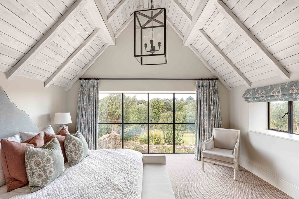 bedroom with apex ceiling cladded in whitewash wood, seagrass carpets and crittal windows overlooking the garden. A large comfy looking bed features with kitsch patterned cushions and matching curtains and blind. An armchair is to the right of the window. 