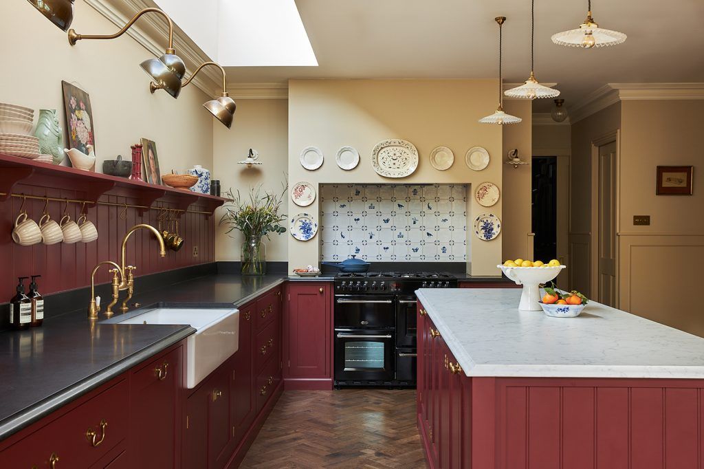 a stylish country-style kitchen with burgundy cabinetry, dark wooden parquet flooring, a kitchen island with marble countertop and cream walls. In the centre a range cooker features with a tiled backsplash and decorative plates.