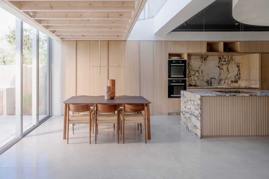 contemporary kitchen/ diner with concrete flooring, light wooden kitchen units and ceiling beams. Off centre is a dark wooden dining table with six wooden chairs, to the right is a waterfall marble island with matching splashback. 