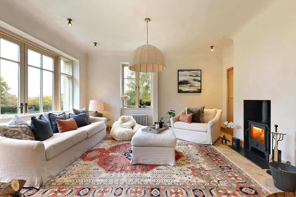 a cream coloured lounge with a lit wood burning stove, large sofa and comfy looking armchair with matching ottoman, set on a large patterned red rug. A large round pendant light hangs above in the centre. The room has french doors to the left and a window on the adjacent wall. 