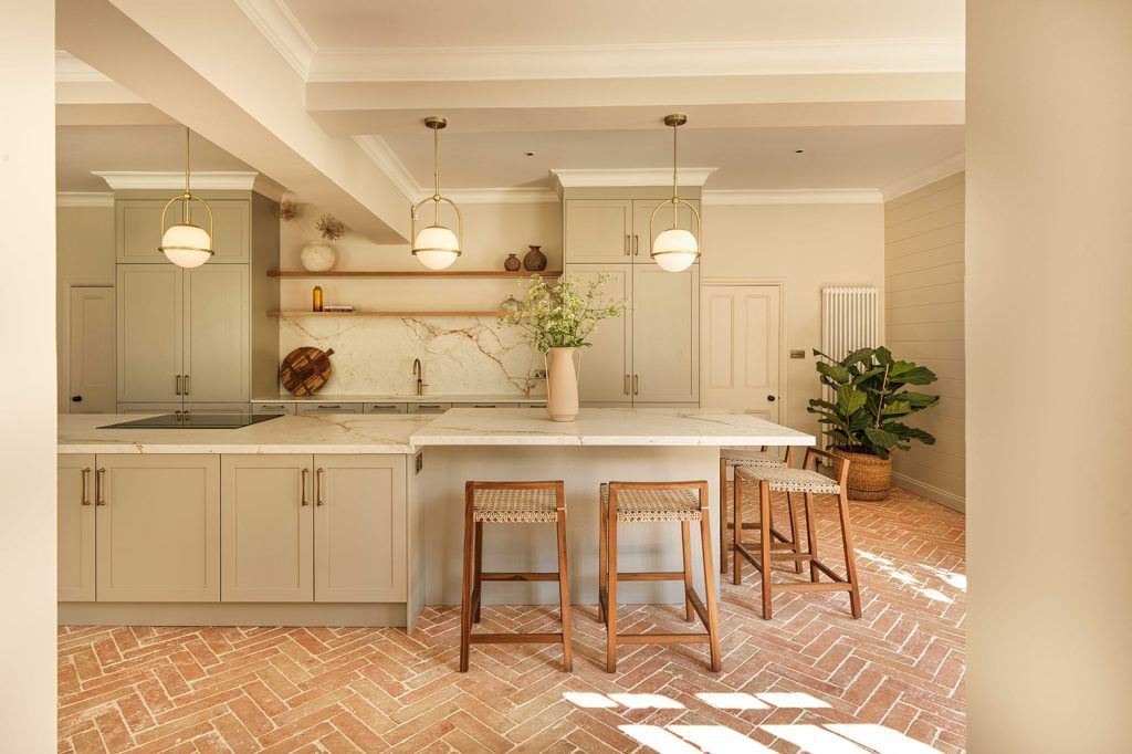 modern kitchen with herringbone brick flooring, a large kitchen island with a marble countertop, integrated hob and four wooden bar stools, three pendant lights hang above, a fiddle leaf fig features. 