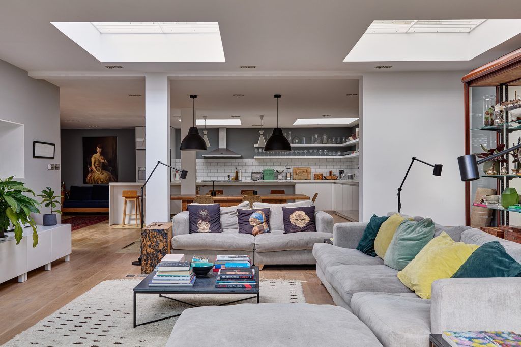 a modern living room with two skylights, two grey sofas, light wooden flooring and a cream rug. In the background you cna see a modern kitchen with a large island, open shelving and wooden bar stools. 