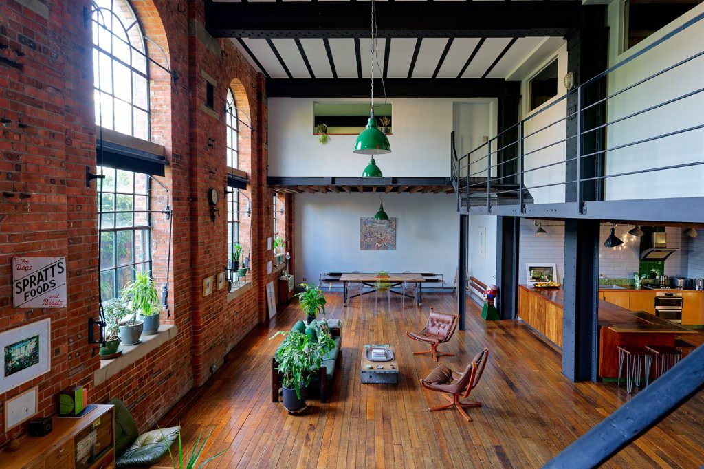 an industrial loft, with crittal arched windows, exposed brick walls, a metal mezzanine staircase and wooden flooring. The layout is open plan with a living room, dining room and kitchen in shot. The flooring is wooden and plants are scattered around the space. 
