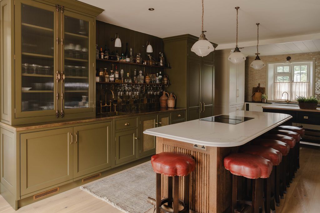 an earthy toned kitchen with olive green cabinetry, a dark wood kitchen island with seven barstools with red leather tops. The island has a marble countertop with an integrated hob and three hanging pendant lights above. In the back of the shot you can see a butchers block island and window. The floor is light wood. 