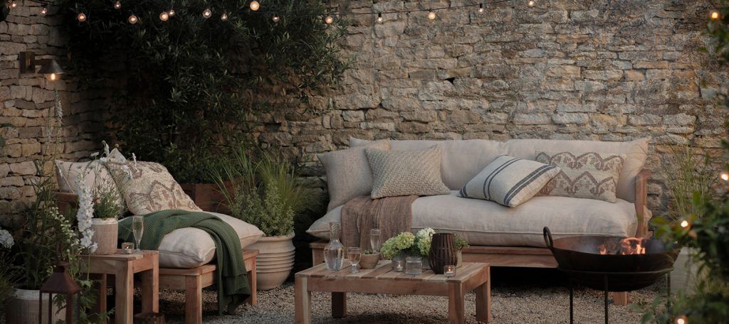 a moody twilight shot of a sofa and chair with comfy cushions outside within a walled garden with stone walls. There is a fire pit with flames visible and greenery throughout the image.