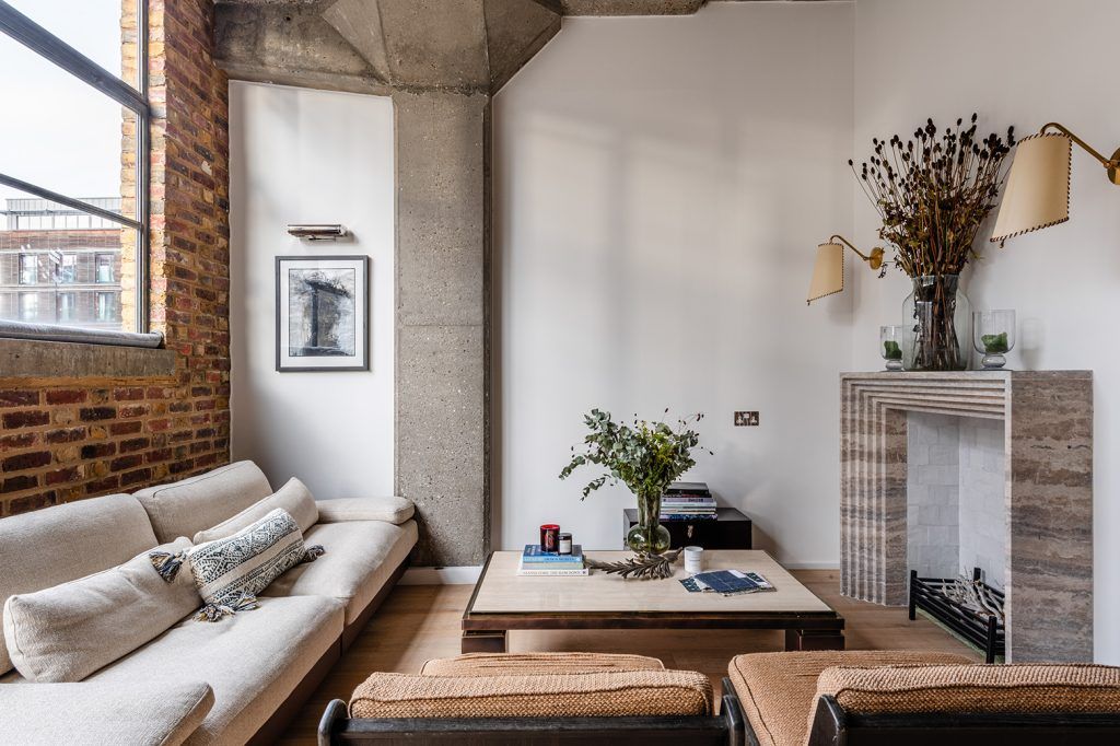 a living room with one exposed brick wall, concrete beam and light wooden flooring. The space features a cream sofa, two accent chairs, a marble fireplace flanked by two wall-lights and dried flowers atop. 