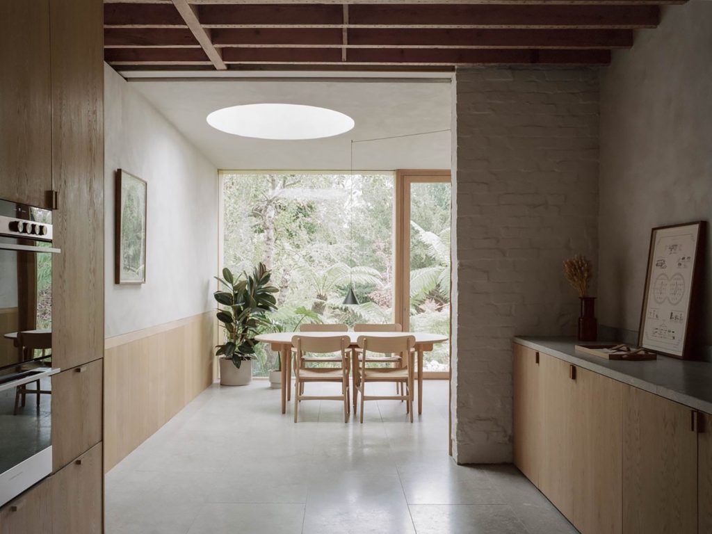 a view from a kitchen into a dining space with a scandi-style table and four chairs. A rubber plant is on the left hand side, with a large glass window showing a tropical gree garden. 