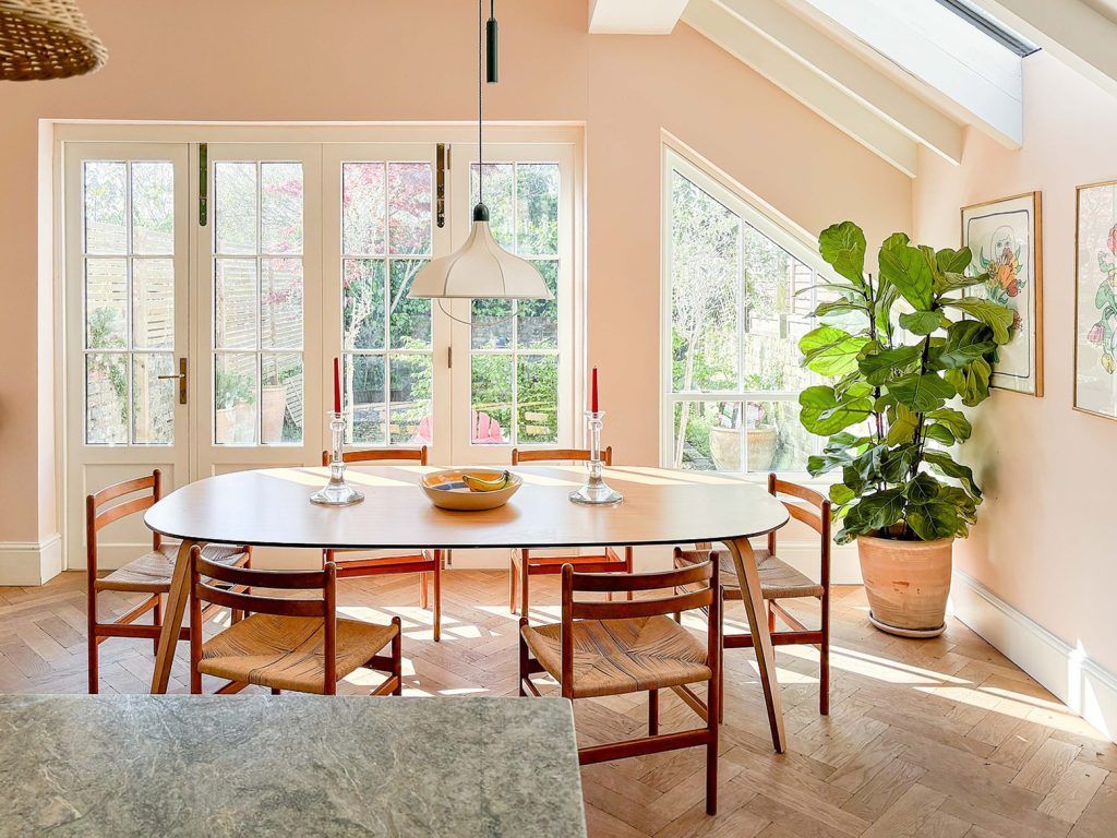 a modern dining room decorated in a soft pink hue with a large oval dining table and six chairs. To the right there is a large fiddle leaf fig plant. French doors lead to the garden. 