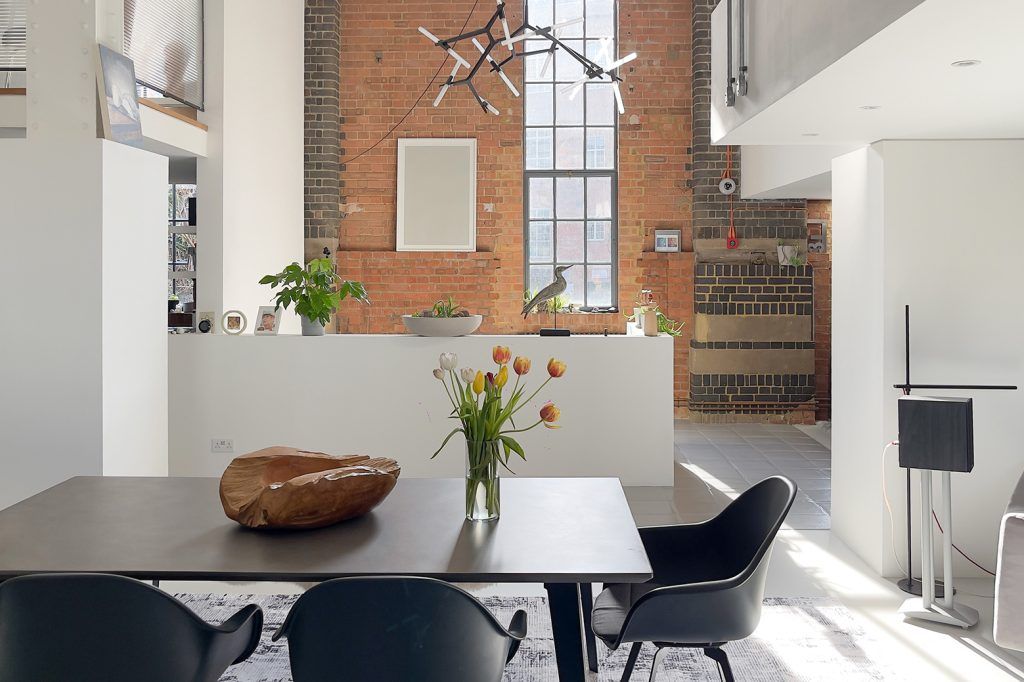 a contemporary loft space set across multiple levels dived by white walls. There is a tall narrow crittal window letting in natural light. The forefront of the image shows a dining table with three plastic eames chairs. On the table is a sculptural piece of wood and tulips in a glass vase. 