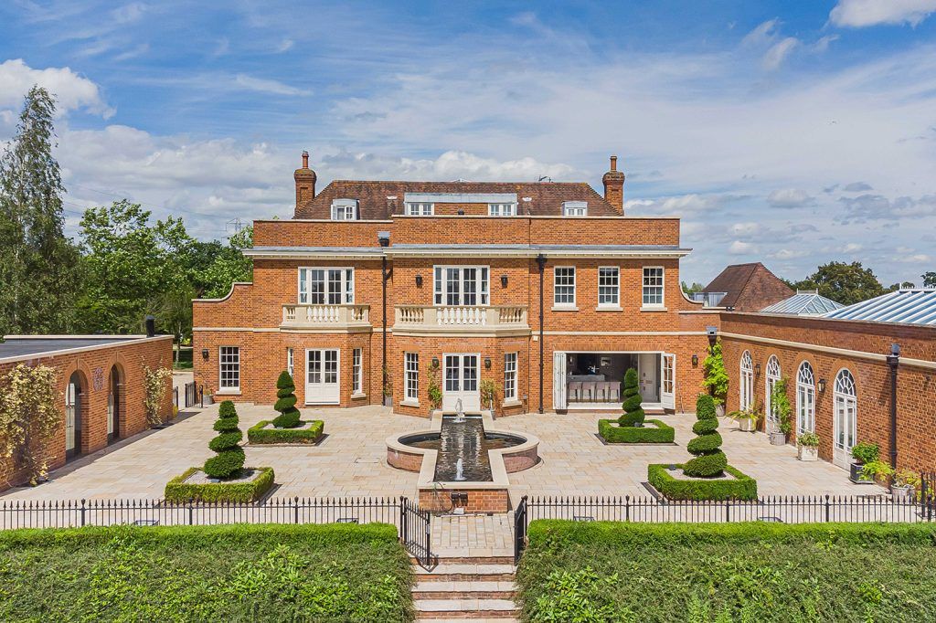 a grand red brick manor house with four sculptural trees, an ornamental pond and tpaved patio. The house is Georgian in style and features sash windows, arched door ways and two balconies. 