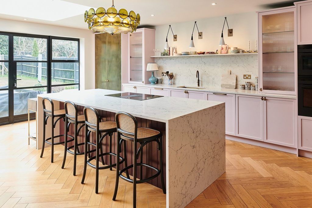 a pastel coloured pink kitchen with a marble waterfall kitchen island with a hob. There is a statement green glass chandelier, parquet flooring and crittal doors to the left of the frame.
