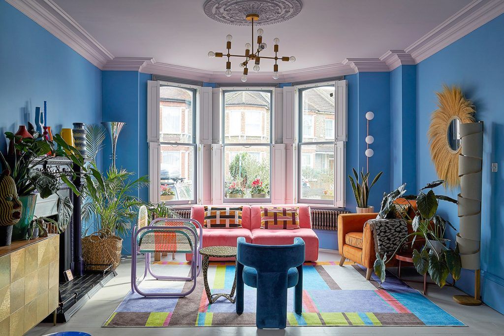 a living room in a Victorian house with bright blue walls, a statement pink sofa, and an array of chairs in different styles and colours, all set on a multi coloured rug.